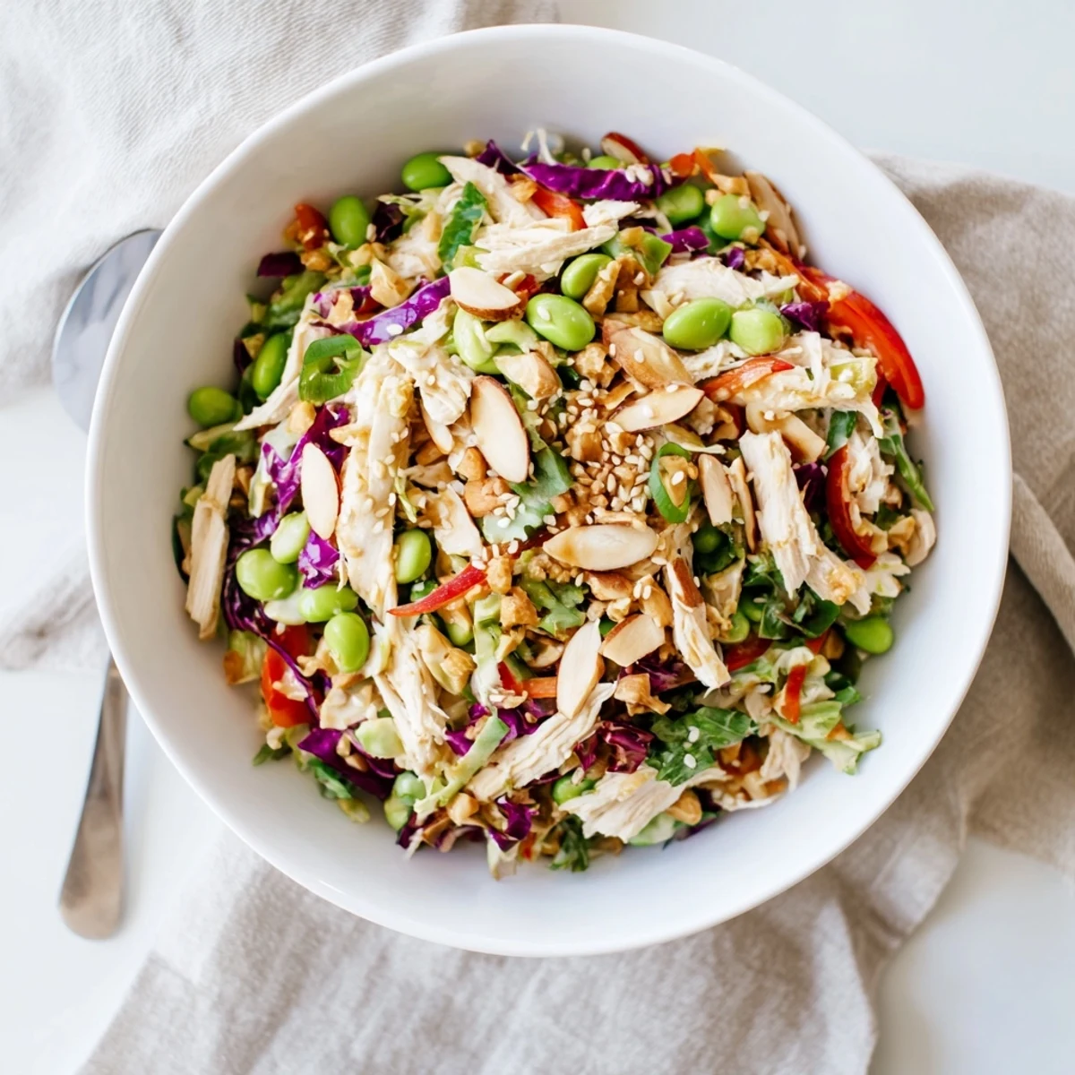 A vibrant Edamame Crunch Chicken Salad in a white bowl features shredded chicken, purple cabbage, carrots, and edamame tossed in a zesty ginger dressing.