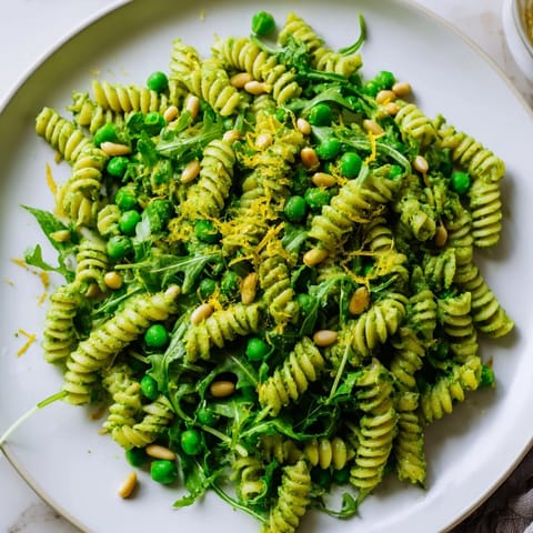 A close-up of Spring Green Pesto Pasta Salad with toasted pine nuts, fresh lemon zest, and crumbled feta cheese on a rustic wooden table.