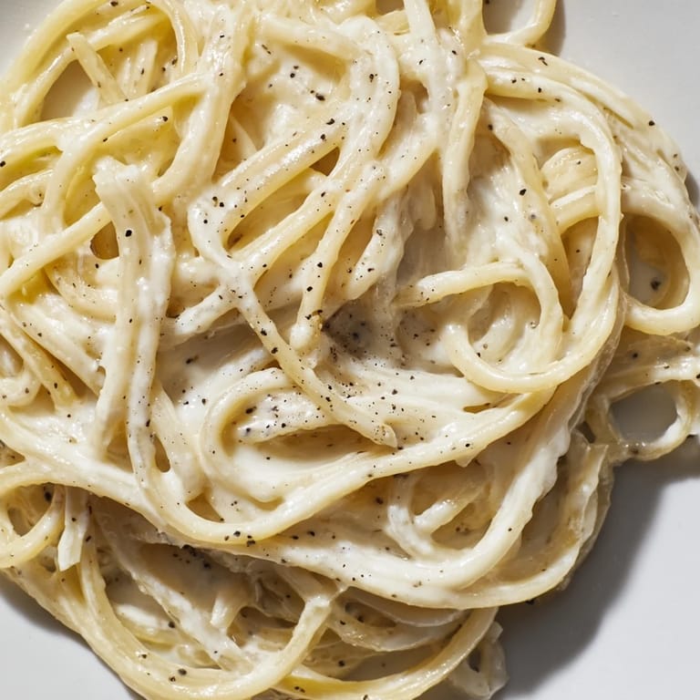 A close-up of spaghetti Cacio e Pepe showing silky cheese sauce clinging to each strand, with cracked pepper on top.